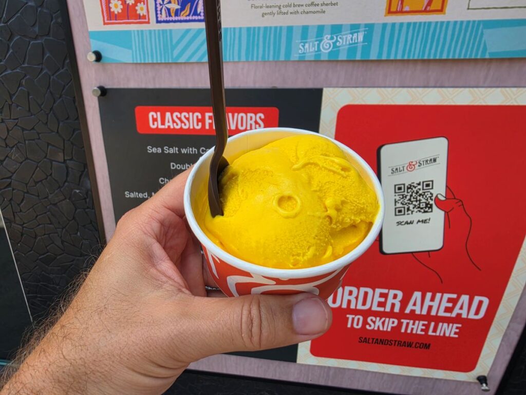 Person holding a cup of bright yellow ice cream with a brown spoon in front of a Salt & Straw signboard.