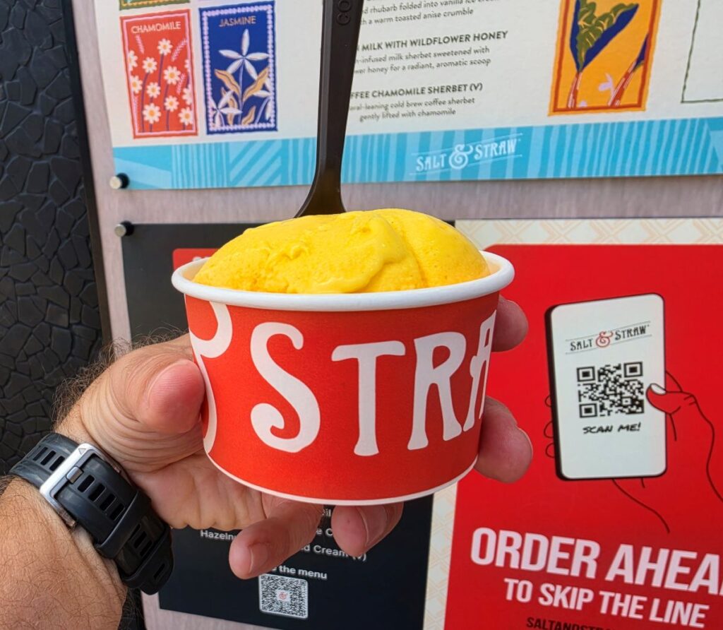 Hand holding a red Salt & StrawDisney Springs cup with a yellow scoop of ice cream, menu board in the background, wristwatch visible on the wrist.