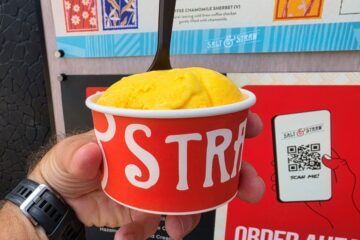 Hand holding a red Salt & StrawDisney Springs cup with a yellow scoop of ice cream, menu board in the background, wristwatch visible on the wrist.