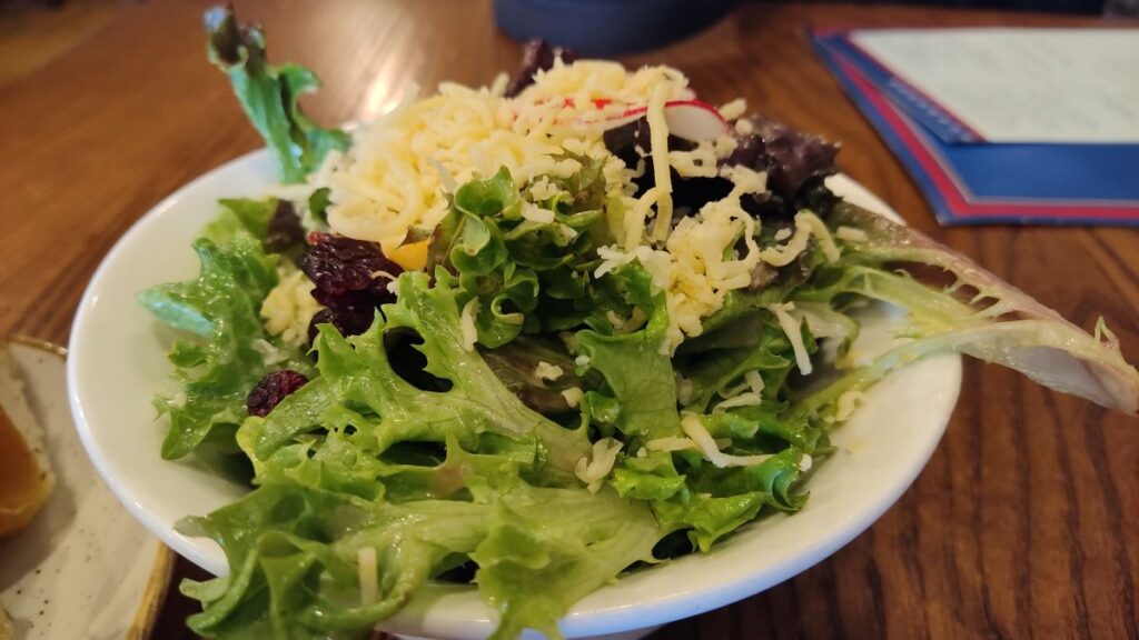 Bowl of mixed green salad with shredded cheese on top. A light vinaigrette appears to glisten on the greens. Eaten during out Liberty Tree Tavern review