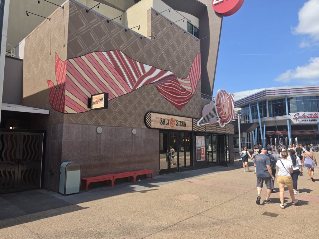 Facade of Salt & Straw Disney Springs shop with a wavy pink striped mural on a brown patterned wall and pedestrians walking by.