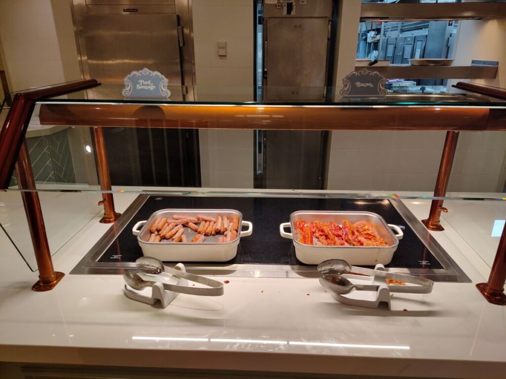 Buffet station with two metal trays of sausage links and bacon, warming on an induction surface, tongs resting nearby.