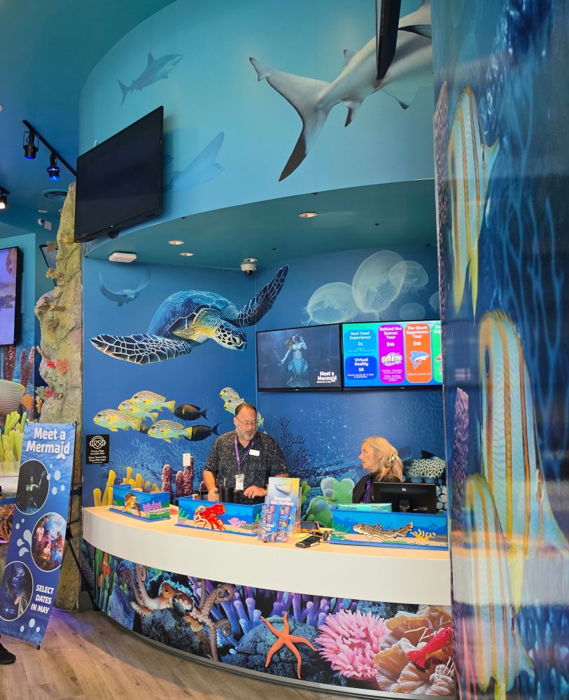 A man and a woman stand behind a curved, ocean-themed reception desk with colorful coral murals and screens on the wall behind them.
