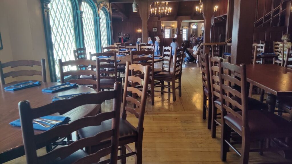 Restaurant dining room with wooden chairs and tables set with blue napkins, warm lighting, and tall arched windows along the left wall; people seated in the distance.