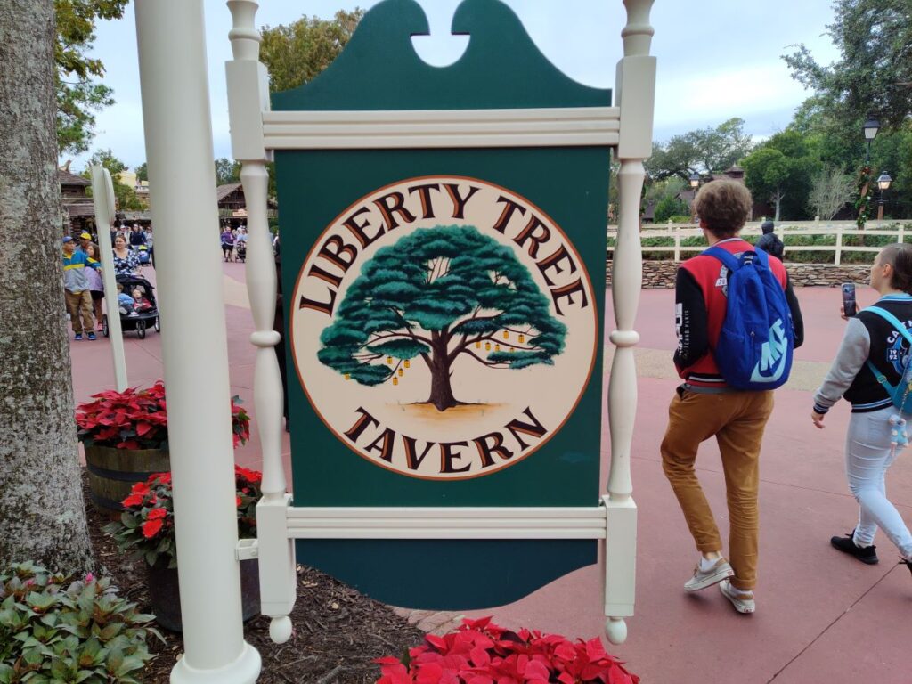 Sign reading Liberty Tree Tavern with a circular emblem of a large tree, framed by cream posts, in an outdoor park walkway with people strolling in the background.