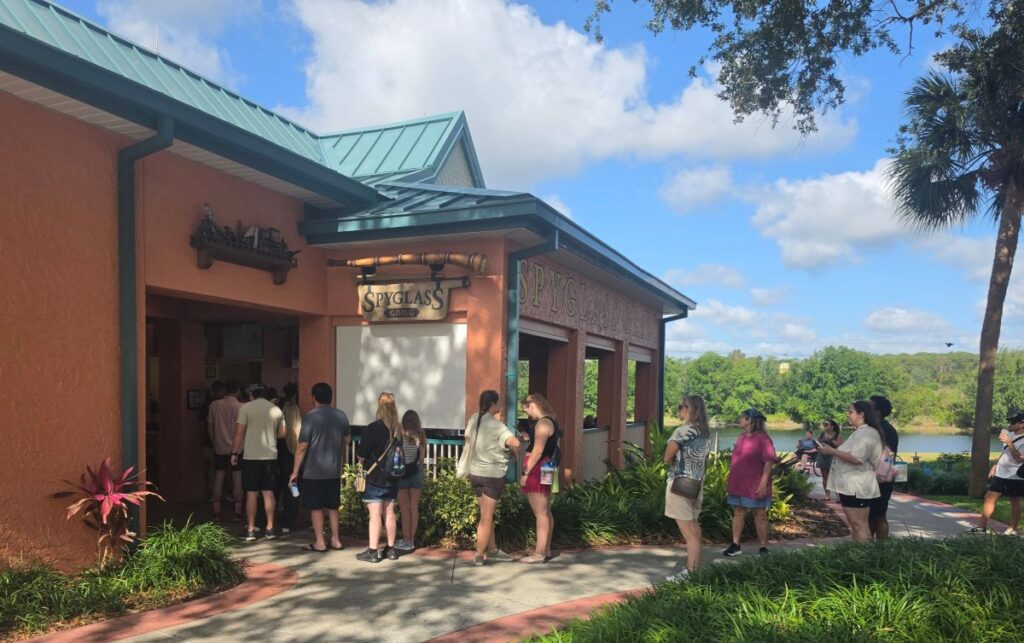 People line up at the Spyglass Grill counter outside a peach-colored building with a teal roof on a sunny day.