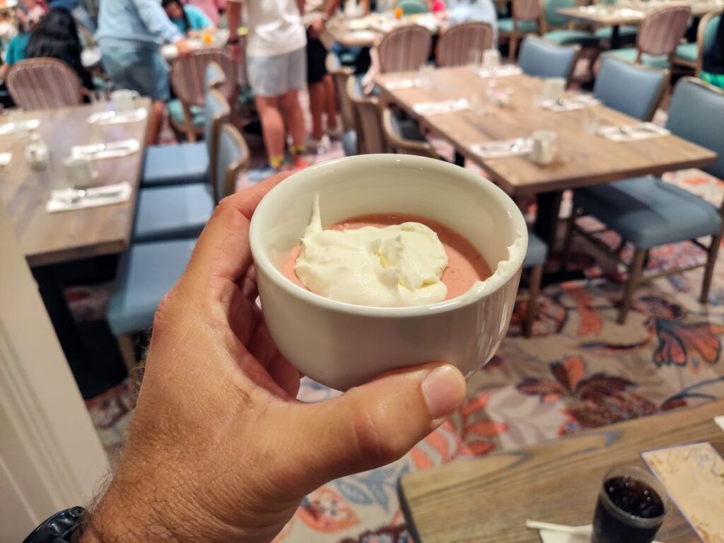 Hand holding a white bowl with pink dessert topped with whipped cream in a busy restaurant.