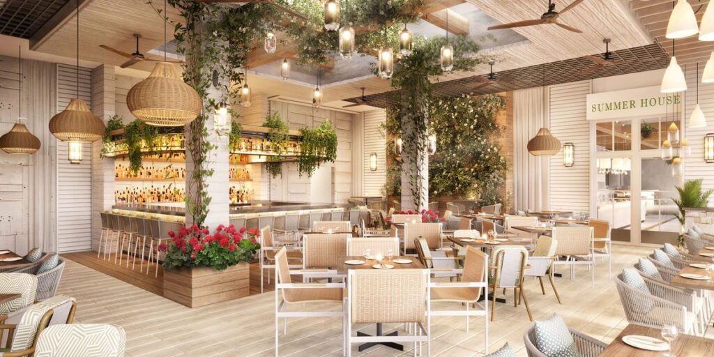 Bright tropical restaurant interior with a long bar, wicker chairs, hanging plants, and warm pendant lights.