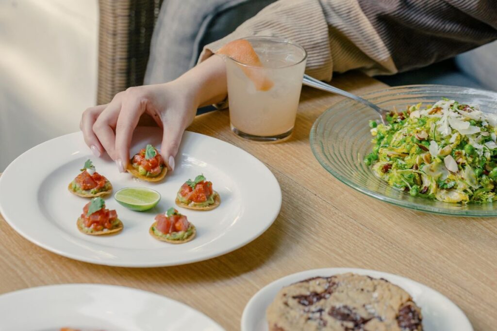 Hand reaching to pick up a bite-sized cracker topped with tomato salsa on a white plate, lime wedge in the center, with a cocktail, a green salad, and a round dessert on a wooden table.