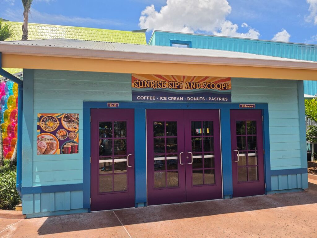 Exterior of a bright turquoise building with purple doors labeled Sunrise Sips and Scoops, offering coffee, ice cream, donuts, and pastries in a sunny setting.