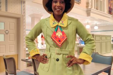 Smiling woman posing in a retro lime-green blazer and matching pants, yellow cuffs, and a straw hat in a bright dining area or cafe with pastel decor.