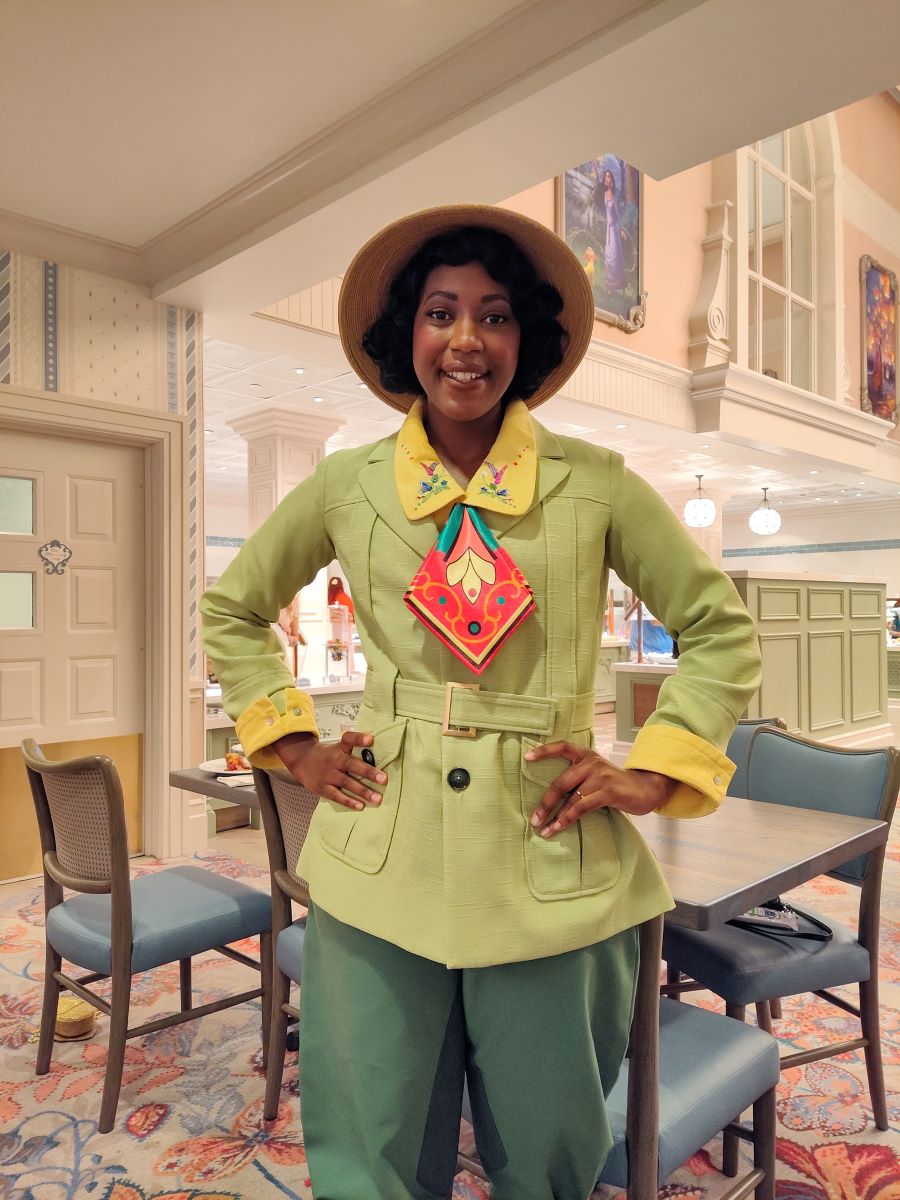 Smiling woman posing in a retro lime-green blazer and matching pants, yellow cuffs, and a straw hat in a bright dining area or cafe with pastel decor.