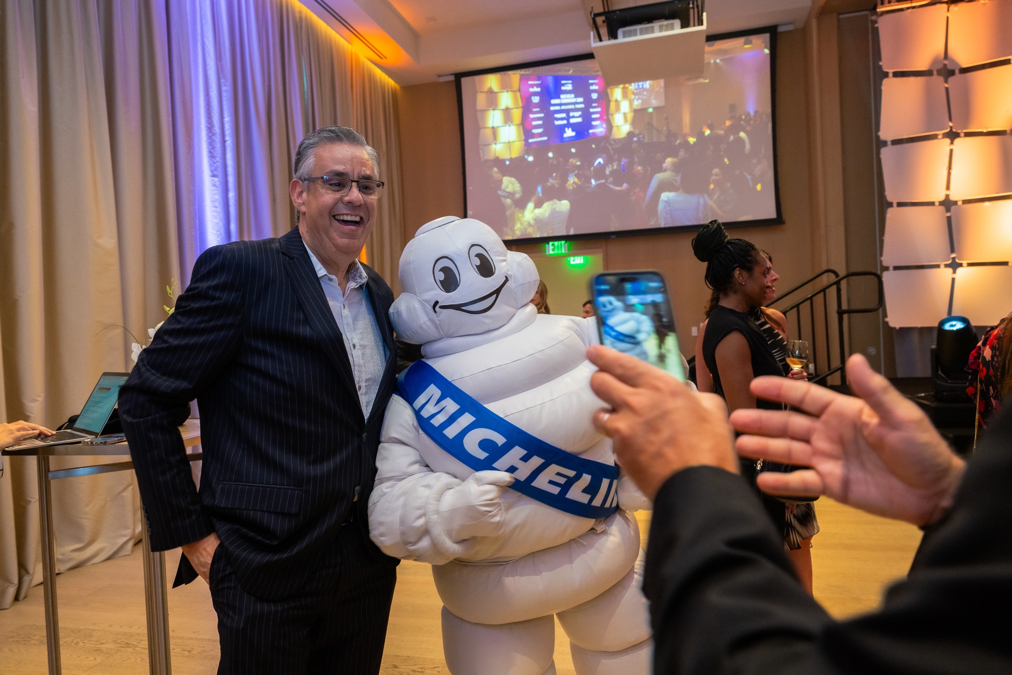 Smiling man in a dark pinstripe suit posing with a life-size Michelin Man mascot at a event, with a projector screen and audience in the background for Victoria & Albert’s Michelin star