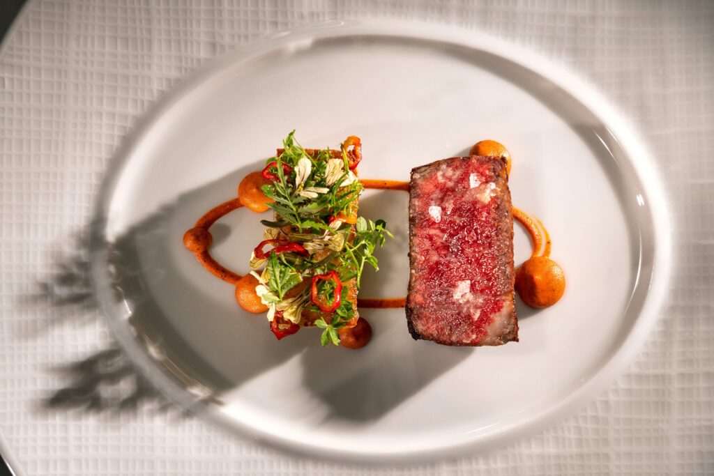Seared steak with a pink center, garnished with herbs and microgreens, arranged on a white plate with orange sauce dots around it