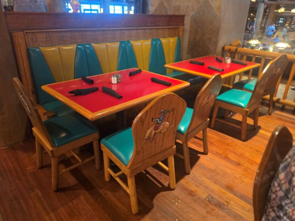Restaurant booth with red tabletops, teal/yellow upholstered benches, and wooden chairs featuring a cartoon dog on the back at Whispering Canyon Cafe Dinner