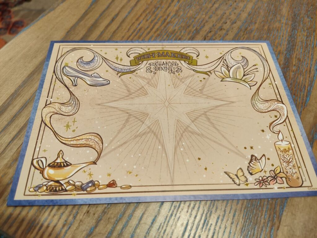 Decorative card with a central starburst and gold decorative border showing a genie lamp, coins, flowers, and butterflies on a wooden table surface.
