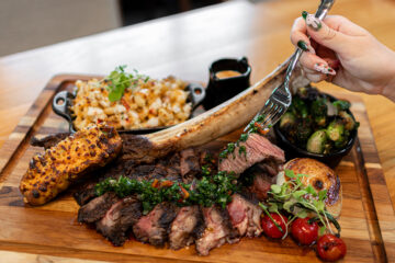 A wooden serving platter with a sliced 34-ounce Tomahawk rib-eye steak as a hand lifts a medium-rare piece of steak with a fork. The feast includes side dishes of lobster mac and cheese, charred Brussels sprouts, a loaded baked potato, grilled onion, and cherry tomatoes for Father's Day Universal CityWalk Orlando