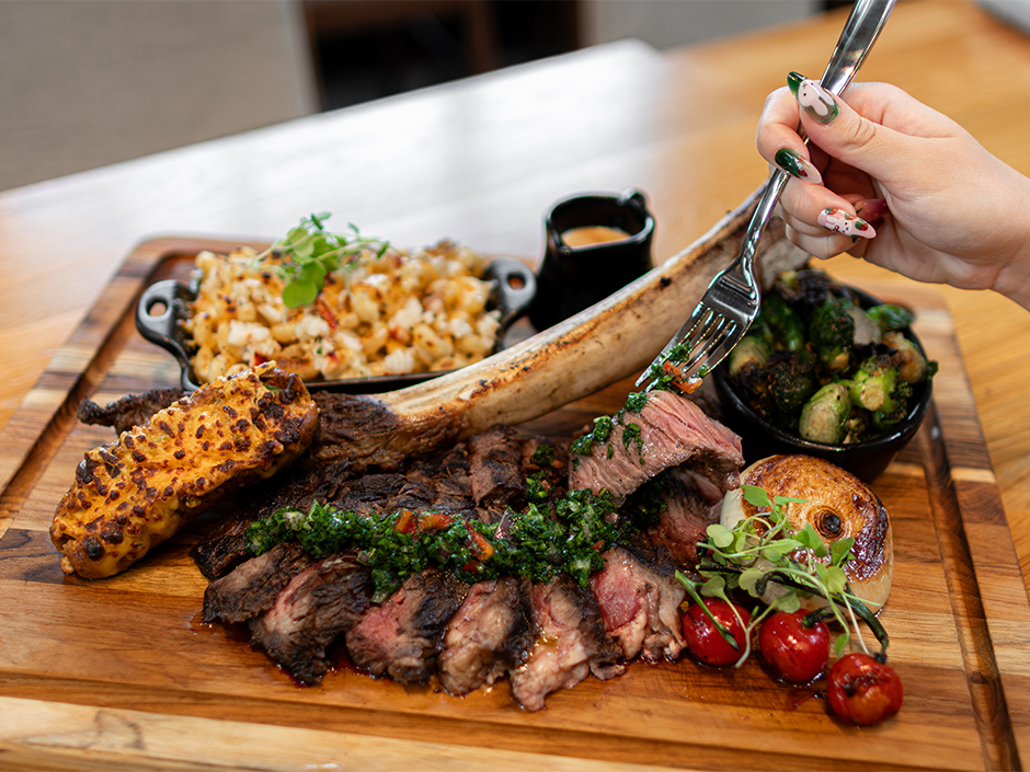 A wooden serving platter with a sliced 34-ounce Tomahawk rib-eye steak as a hand lifts a medium-rare piece of steak with a fork. The feast includes side dishes of lobster mac and cheese, charred Brussels sprouts, a loaded baked potato, grilled onion, and cherry tomatoes for Father's Day Universal CityWalk Orlando