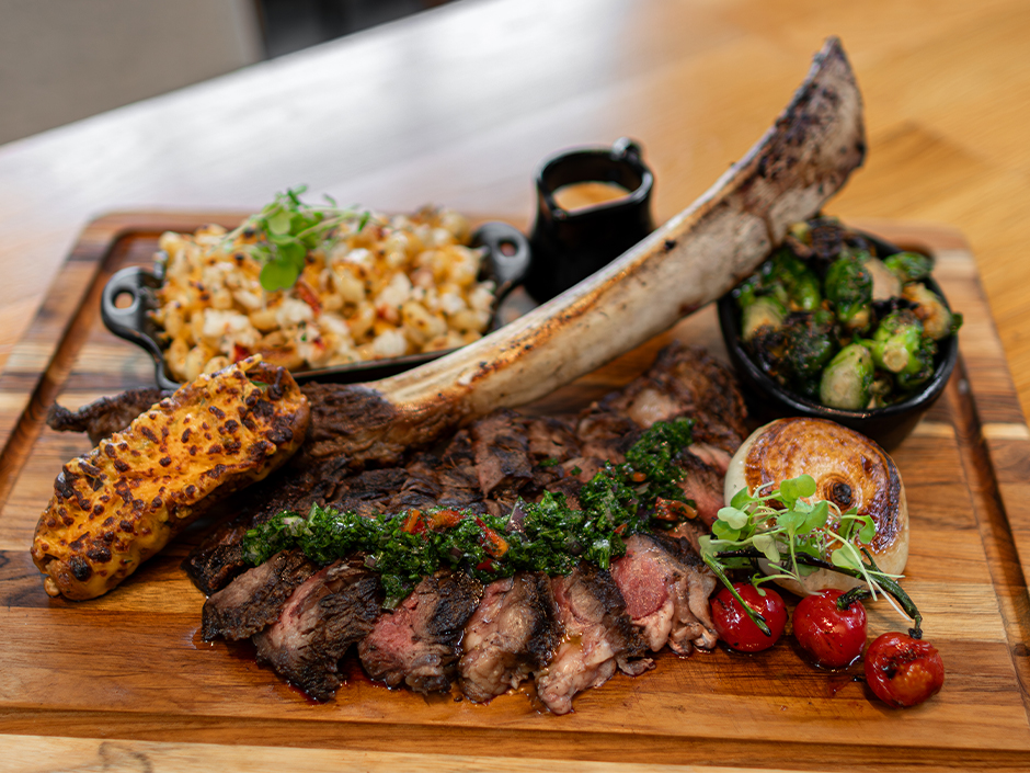 A wooden serving board featuring a massive sliced 34-ounce Tomahawk rib-eye steak with its long bone extending across the board. The steak is topped with green chimichurri sauce and accompanied by sides of lobster macaroni and cheese, roasted Brussels sprouts, a twice-baked potato, a grilled onion half, and roasted cherry tomatoes.