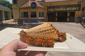 A close-up view of a hand holding a food tray with the "Pulled Pork Mac & Cheese Cone" from SeaWorld Orlando. A golden waffle cone is packed with savory BBQ pulled pork and set beside a heavily charred, seasoned corn rib. The background frames the vibrant "Summer Bites" kiosk and the adjacent Voyager's Smokehouse under the afternoon sun.