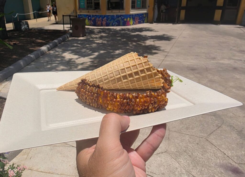 A close-up shot of a paper plate held by a hand, focusing on the curved, caramelized corn rib lying in front of a waffle cone filled with BBQ pulled pork. The scene is captured outdoors on a sunny day, with the shadows of nearby trees stretching across the concrete path toward the Voyager's Smokehouse building.