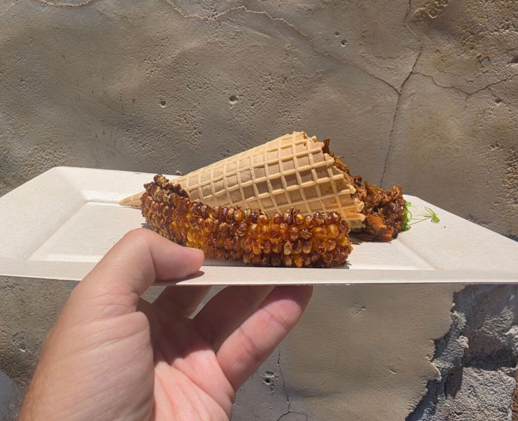 A close-up, tight shot of a snack tray held up against a textured, light grey stucco wall. The plate contains a BBQ Pork Mac Cone, garnished with microgreens, and overflowing with dark, saucy pulled pork garnished with microgreens, nestled next to a heavily roasted and seasoned corn rib.