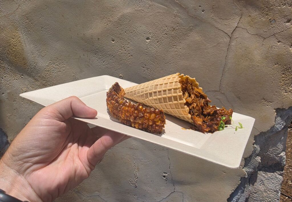 An angled, first-person shot of a snack tray held in front of a sunlit, weathered concrete wall. The tray holds a waffle cone tipped on its side, spilling out rich, dark barbecue pulled pork, accompanied by a charred, curved corn rib.
