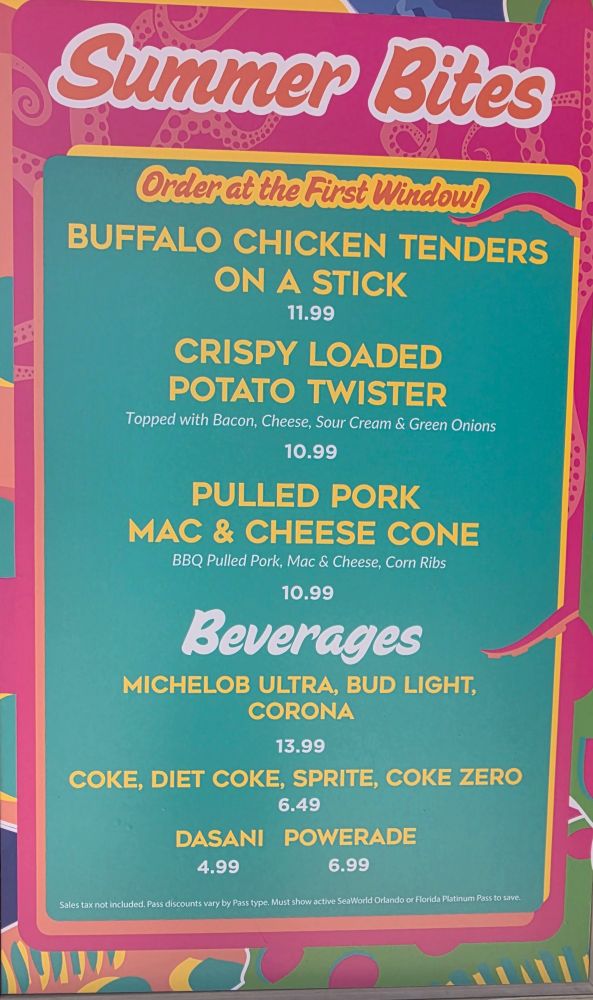 A vertical, close-up shot of the colorful "Summer Bites" outdoor menu sign. The top header reads "Summer Bites" in white cursive over a pink backdrop with a faint octopus tentacle pattern. The main teal-colored section reads "Order at the First Window!" followed by menu items and prices in yellow text: "Buffalo Chicken Tenders on a Stick ($11.99)", "Crispy Loaded Potato Twister ($10.99)", and "Pulled Pork Mac & Cheese Cone ($10.99)" described as having BBQ Pulled Pork, Mac & Cheese, and Corn Ribs. Beverage options listed include beer, sodas, water, and Powerade.