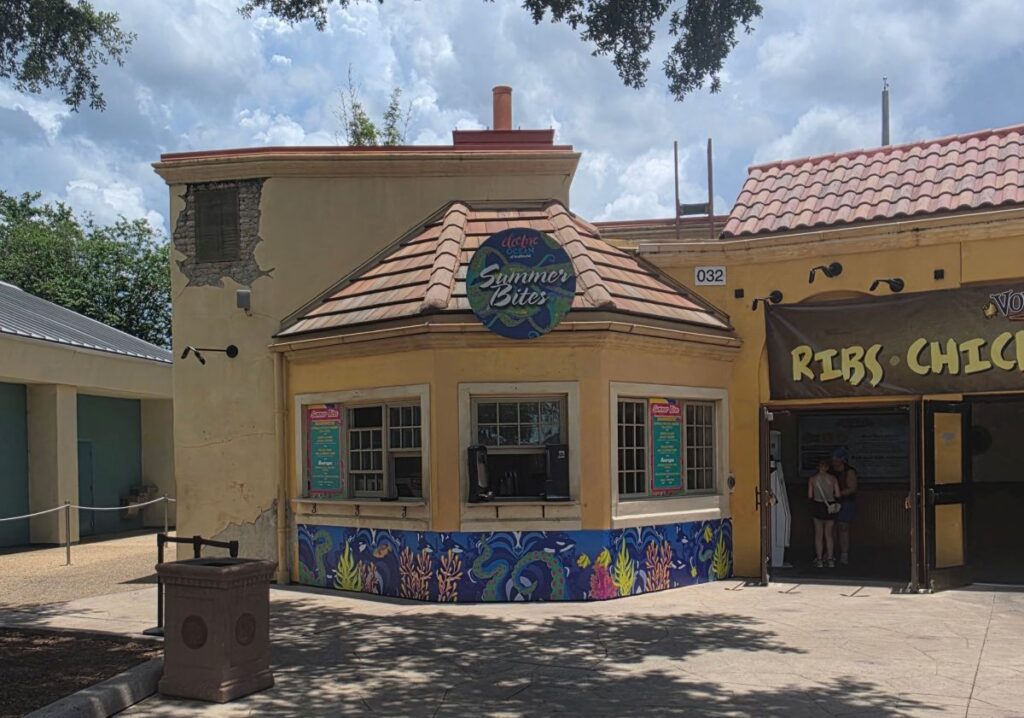 A wide, daytime shot of the "Summer Bites" food kiosk at SeaWorld Orlando - The circular, yellow stucco building features a colorful under-counter mural depicting stylized coral, sea plants, and octopus tentacles. A large circular sign on the tiled roof reads "Electric Ocean Summer Bites." To the right, an open doorway leads into Voyager's Smokehouse beneath a brown banner that lists "Ribs • Chicken • Brisket."