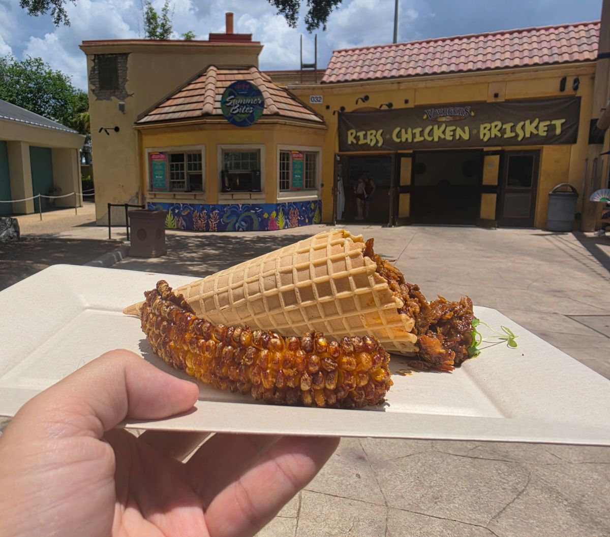 A close-up view of a hand holding a food tray with the "Pulled Pork Mac & Cheese Cone" from SeaWorld Orlando. A golden waffle cone is packed with savory BBQ pulled pork and set beside a heavily charred, seasoned corn rib. The background frames the vibrant "Summer Bites" kiosk and the adjacent Voyager's Smokehouse under the afternoon sun.