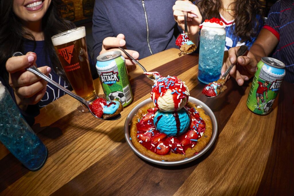 A top-down view of a wooden restaurant table where four people are sharing a large Stars and Stripes Pizookie from BJ's Restaurant & Brewhouse. The dessert features a warm cookie in a metal skillet topped with a scoop of blue ice cream, a scoop of vanilla ice cream, strawberry toppings, and red, white, and blue sprinkles. Each person holds a spoon lifted with a bite of the dessert. The table also holds a pint of BJ's beer, two cans of Grand Attack Cold IPA, and two vibrant blue cocktails topped with whipped cream.