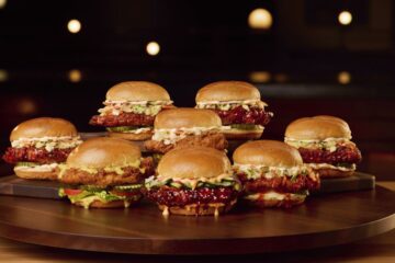 A lineup of eight different BJ's Restaurant & Brewhouse crispy chicken sandwiches arranged on a wooden platter, featuring various sauces, slaws, and toppings on toasted buns.