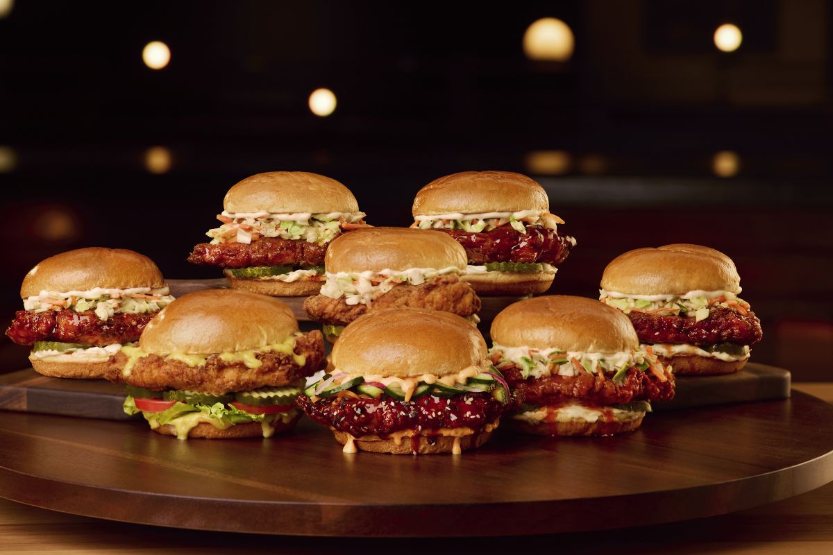 A lineup of eight different BJ's Restaurant & Brewhouse crispy chicken sandwiches arranged on a wooden platter, featuring various sauces, slaws, and toppings on toasted buns.