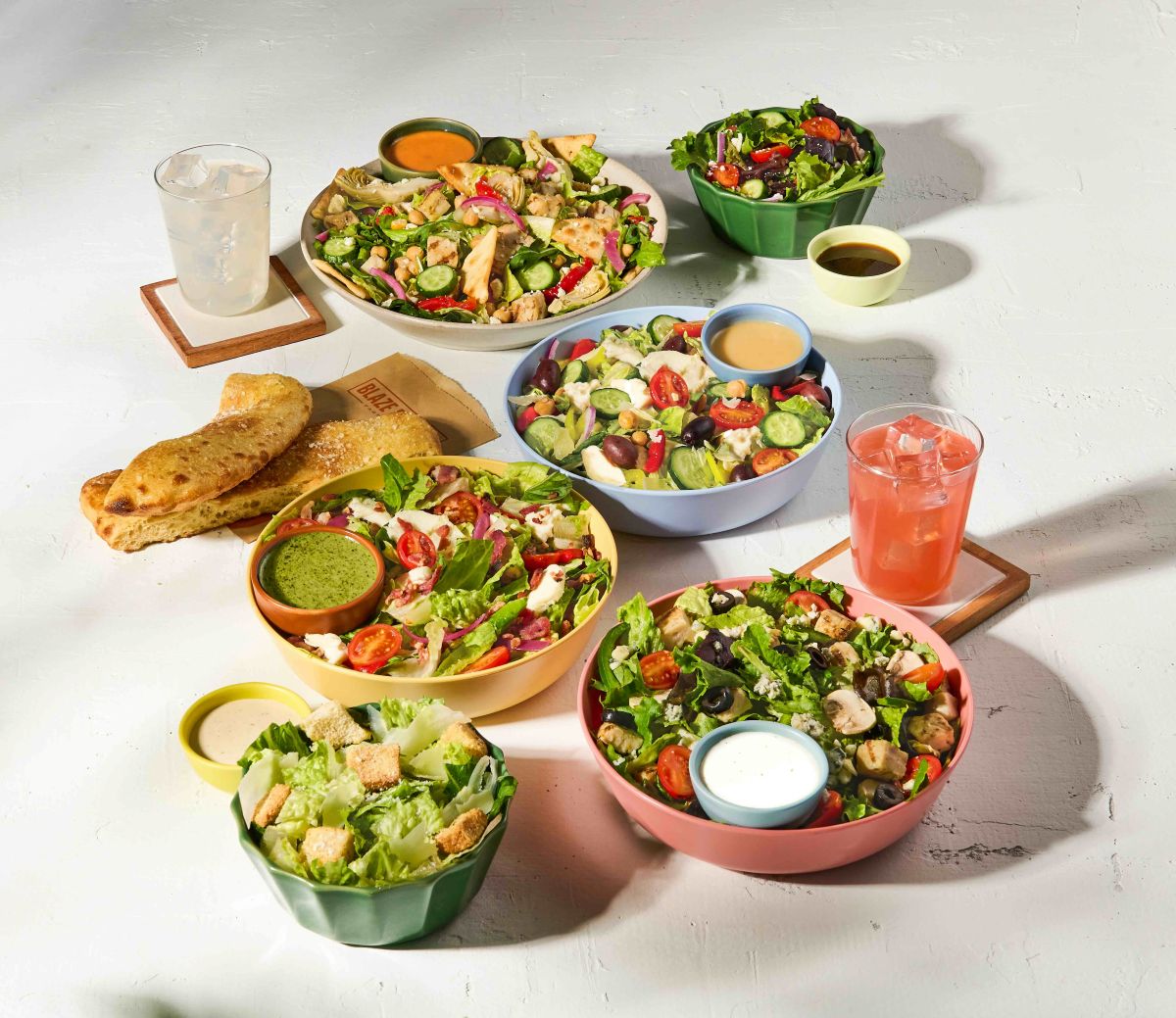 A variety of fresh Blaze Pizza entree salads topped with colorful vegetables, chicken, and dressings, served alongside garlic breadsticks and cold drinks for a summer meal.