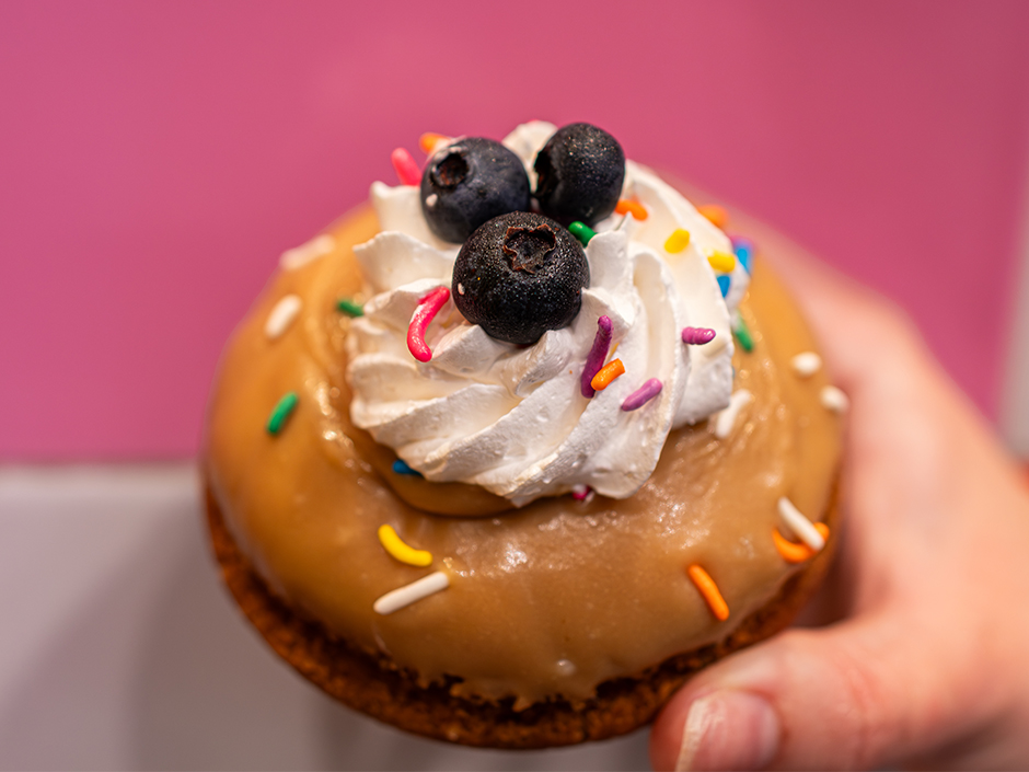 A close-up shot of a hand holding a gourmet yeast doughnut from Voodoo Doughnut against a bright pink background. The doughnut is topped with smooth maple glaze, a swirl of white whipped cream, multi-colored sprinkles, and three fresh blueberries.