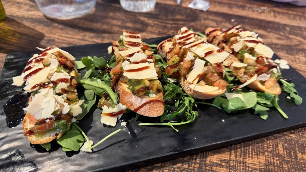Four pieces of crispy toasted bruschetta topped with diced tomatoes, fresh herbs, shaved parmesan cheese, and a rich balsamic drizzle, served over a bed of green arugula on a black platter at Terralina Crafted Italian.
