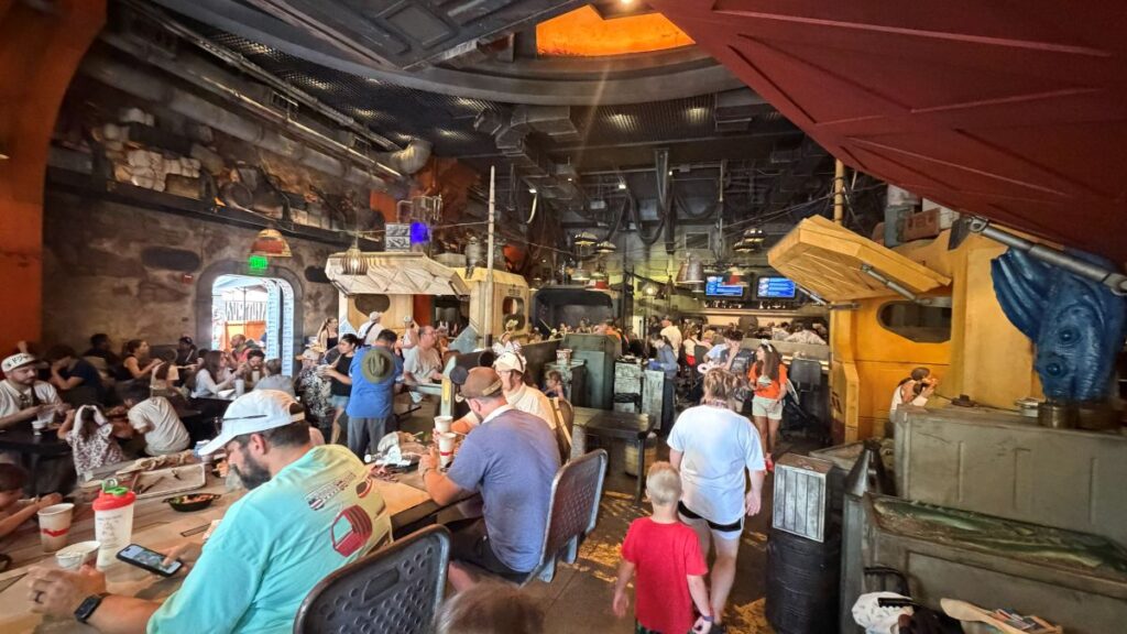The busy, industrial interior of a Docking Bay 7 dining room, styled like a hangar with exposed pipes, cargo crates, overhead lighting, and crowds of people eating at various tables.