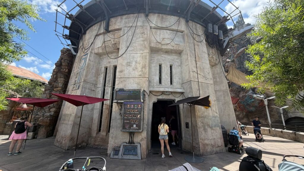 A stone-colored, industrial-style circular building entrance for Docking Bay 7 Food and Cargo under a bright blue sky, featuring wires draped across the exterior and red patio umbrellas nearby.