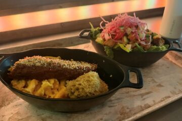 wo Star Wars-themed entrees served in black, oval-shaped dishes on a metal tray, featuring a plate of macaroni and cheese with a seasoned rib on top next to a vibrant chopped salad topped with pink crispy noodles.