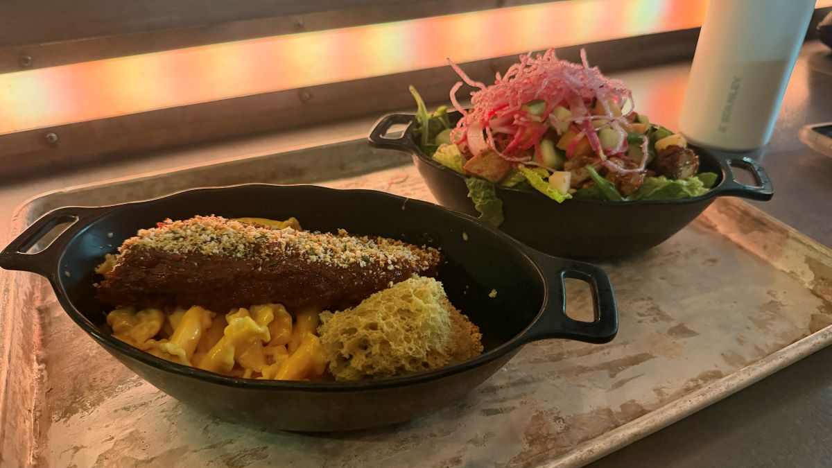 wo Star Wars-themed entrees served in black, oval-shaped dishes on a metal tray, featuring a plate of macaroni and cheese with a seasoned rib on top next to a vibrant chopped salad topped with pink crispy noodles.