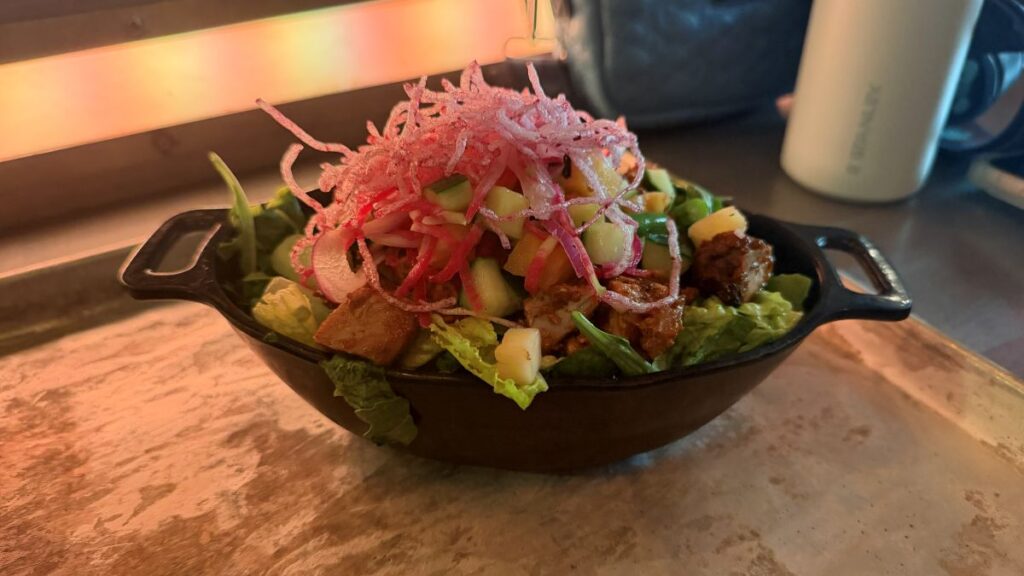 A vibrant Endorian Chicken Tip-Yip Salad served in a black dish, consisting of leafy greens, diced chicken, chopped cucumbers, and mango, topped with a pile of shredded pink crispy noodles.