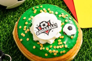 The Everglazed summer soccer donut featuring green icing, gold sprinkles, a dollop of white buttercream, an edible "GOOAAALL!" disc, and a candy soccer ball, styled on green turf next to a referee whistle and penalty cards.