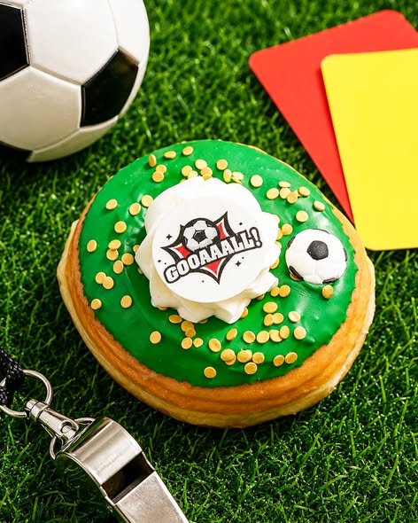 The Everglazed summer soccer donut featuring green icing, gold sprinkles, a dollop of white buttercream, an edible "GOOAAALL!" disc, and a candy soccer ball, styled on green turf next to a referee whistle and penalty cards.