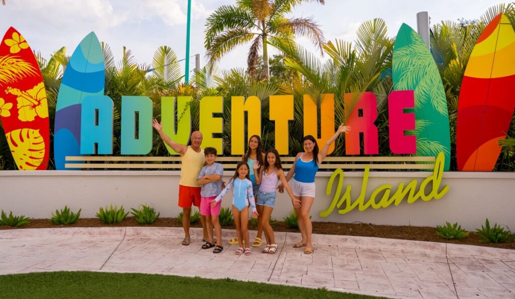 A happy family of six posing and waving in front of the colorful, giant Adventure Island sign surrounded by palm trees and surfboards at AquaGlow Adventure Island Tampa.