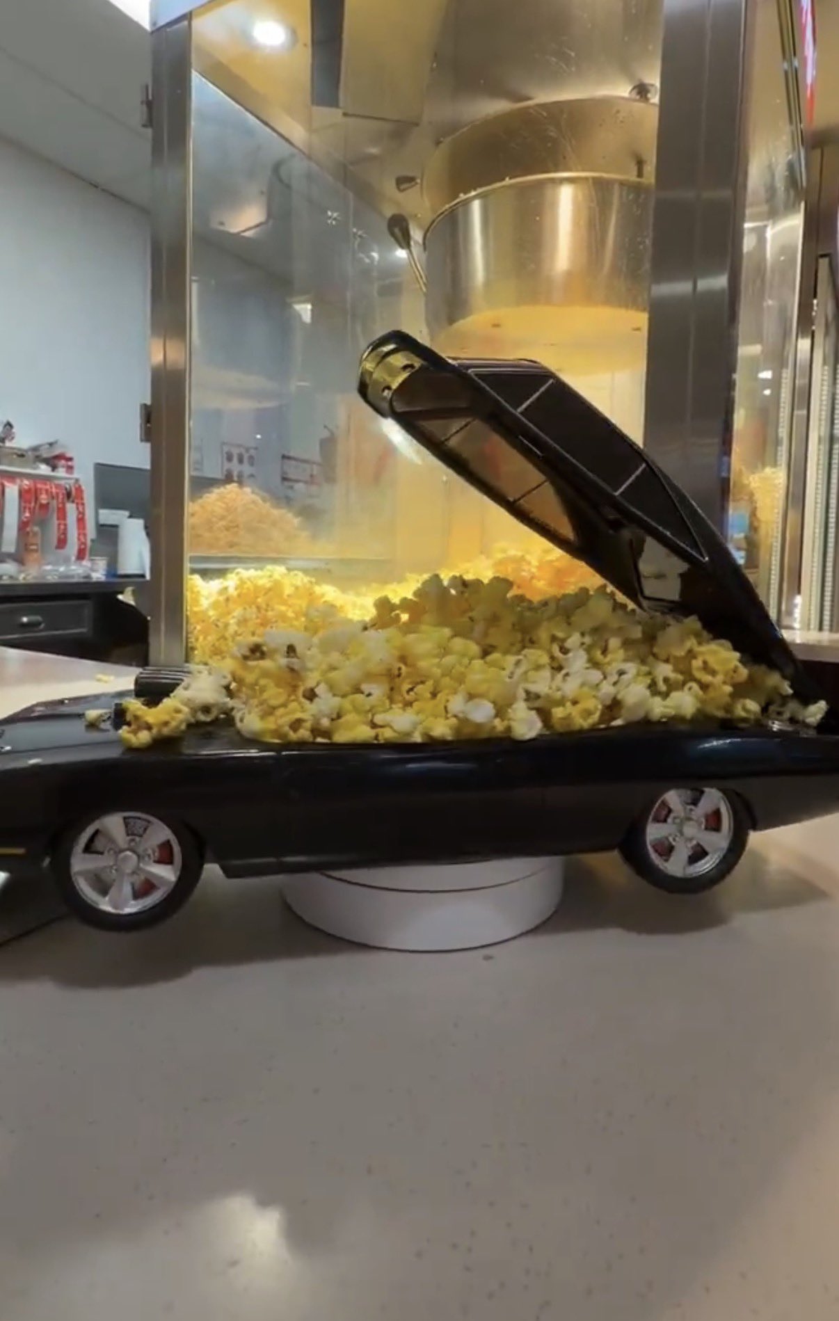 A close-up shot of official Fast & Furious merch at a Cinemark concession stand, showing the black Dodge Charger collectible popcorn bucket with its top opened up and filled to the brim with fresh movie theater popcorn.