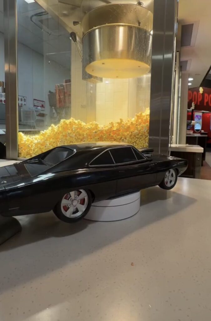 A sleek black Dodge Charger collectible popcorn bucket displayed on a white stand at a movie theater counter, showcasing the detailed design of this exclusive Fast & Furious merch in front of a popcorn machine.