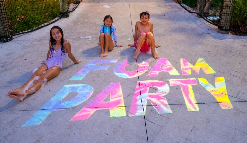 Three smiling kids sitting on a paved walkway around a colorful neon "Foam Party" light projection at AquaGlow Adventure Island Tampa.