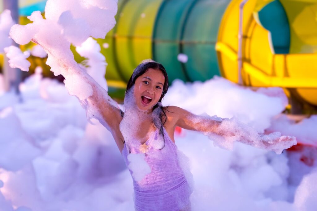 A joyful girl covered in bubbles smiling with her arms open during a foam party near a large water slide at AquaGlow Adventure Island Tampa.