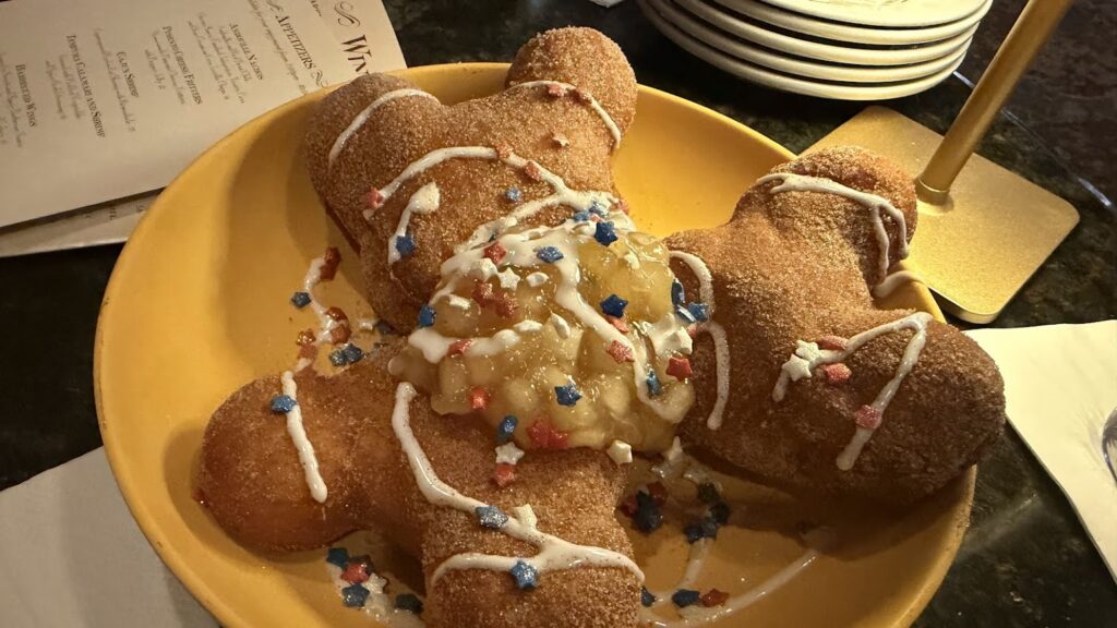 An angled, overhead view of three golden-brown, Mickey-shaped Fourth if July beignets served on a yellow plate. The treats are coated in cinnamon sugar, topped with a spoonful of warm apple compote, crisscrossed with white icing, and finished with red, white, and blue star sprinkles. A dim restaurant tabletop and an appetizer menu are visible in the background.