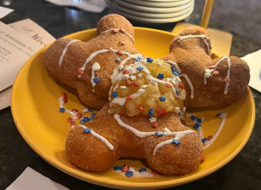 A vibrant yellow plate holds three Mickey-shaped beignets dusted with cinnamon sugar. The pastries are arranged around a central heap of diced apple compote, then drizzled with white icing and scattered with festive red, white, and blue star-shaped sprinkles. A restaurant menu is partially visible in the background.